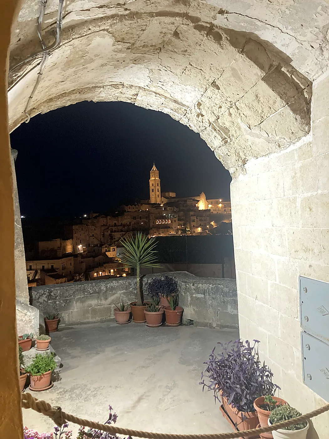 I Sassi di Matera illuminati di notte, con il campanile del Duomo, visti attraverso un arco di pietra da una terrazza fiorita.