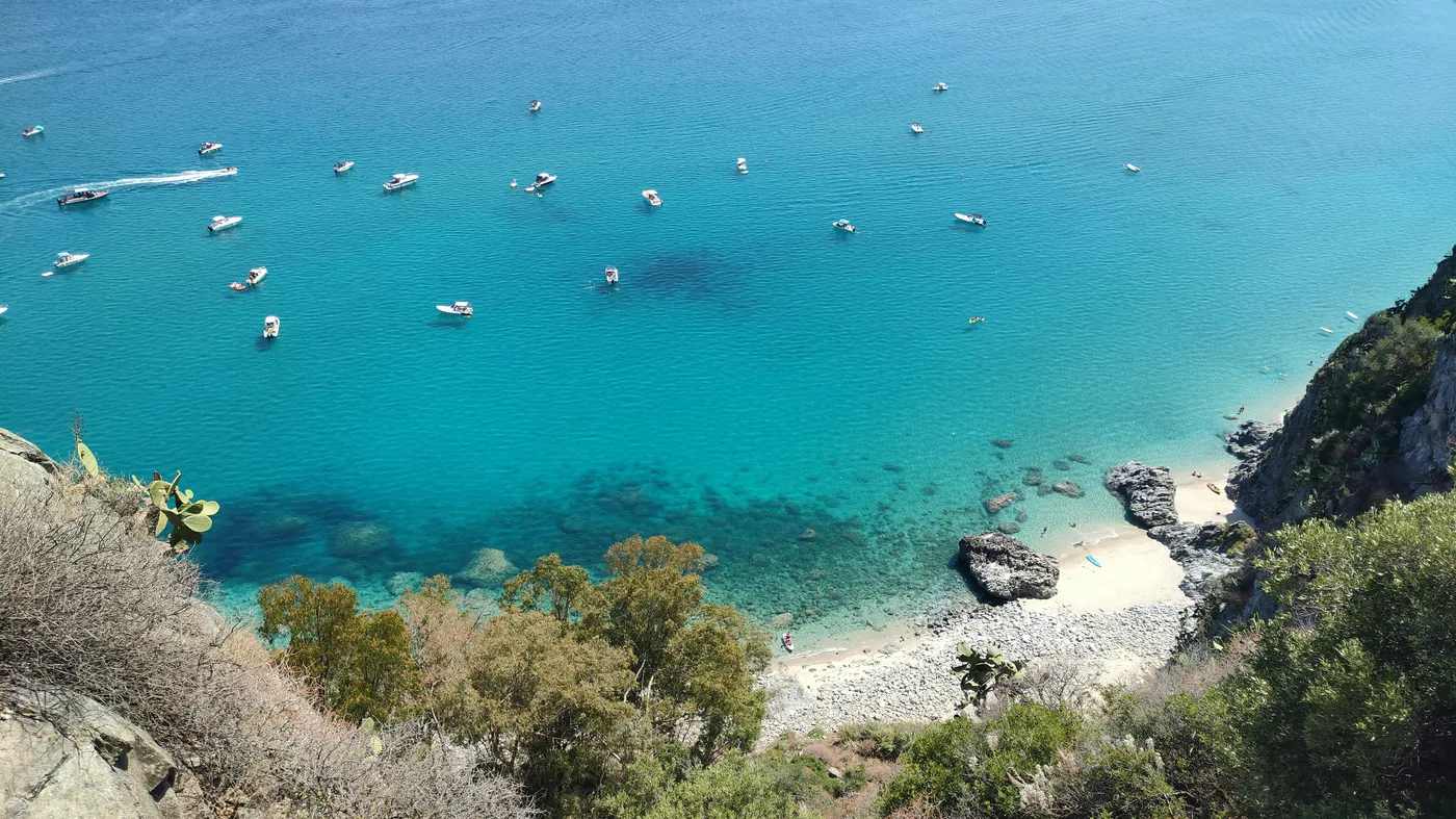 Una cala vista dall'alto, con barche ancorate nell'acqua turchese e una spiaggia di ciottoli chiari
