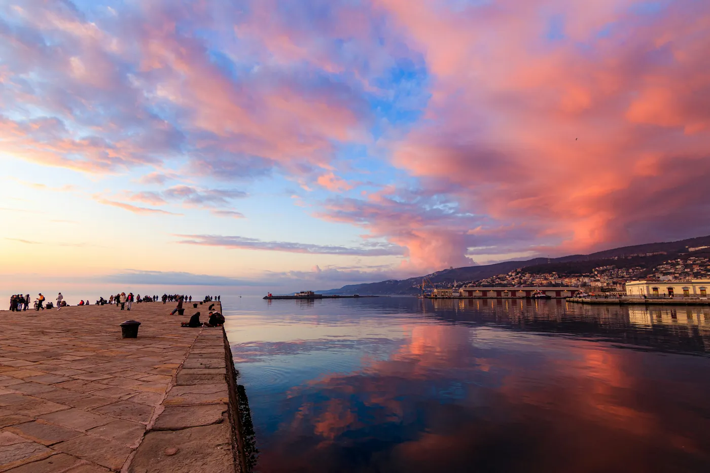 Il molo Audace di Trieste al tramonto, con le nuvole rosa riflesse nel mare e il porto in lontananza