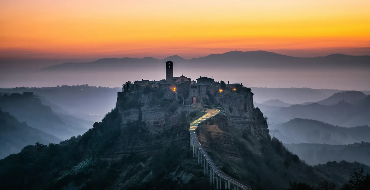 Civita di Bagnoregio sulla sua rupe, con il ponte pedonale illuminato, nella luce arancione dell'alba