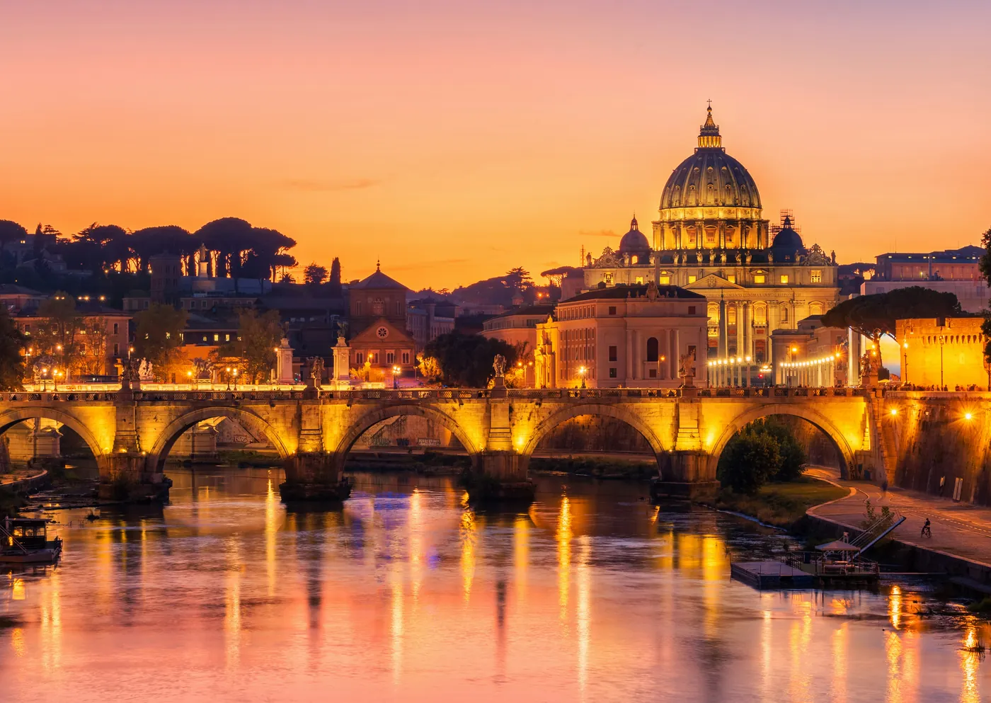 Ponte Sant'Angelo illuminato sul Tevere e la cupola di San Pietro nel cielo arancione della sera
