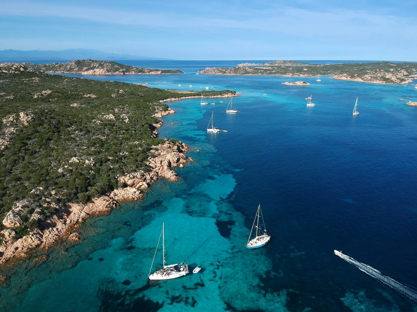 Barche a vela all'ancora davanti a una costa di granito e macchia, in un mare blu e turchese