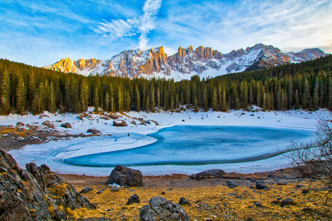 Il lago di Carezza ghiacciato tra gli abeti, con le cime del Latemar illuminate dal sole