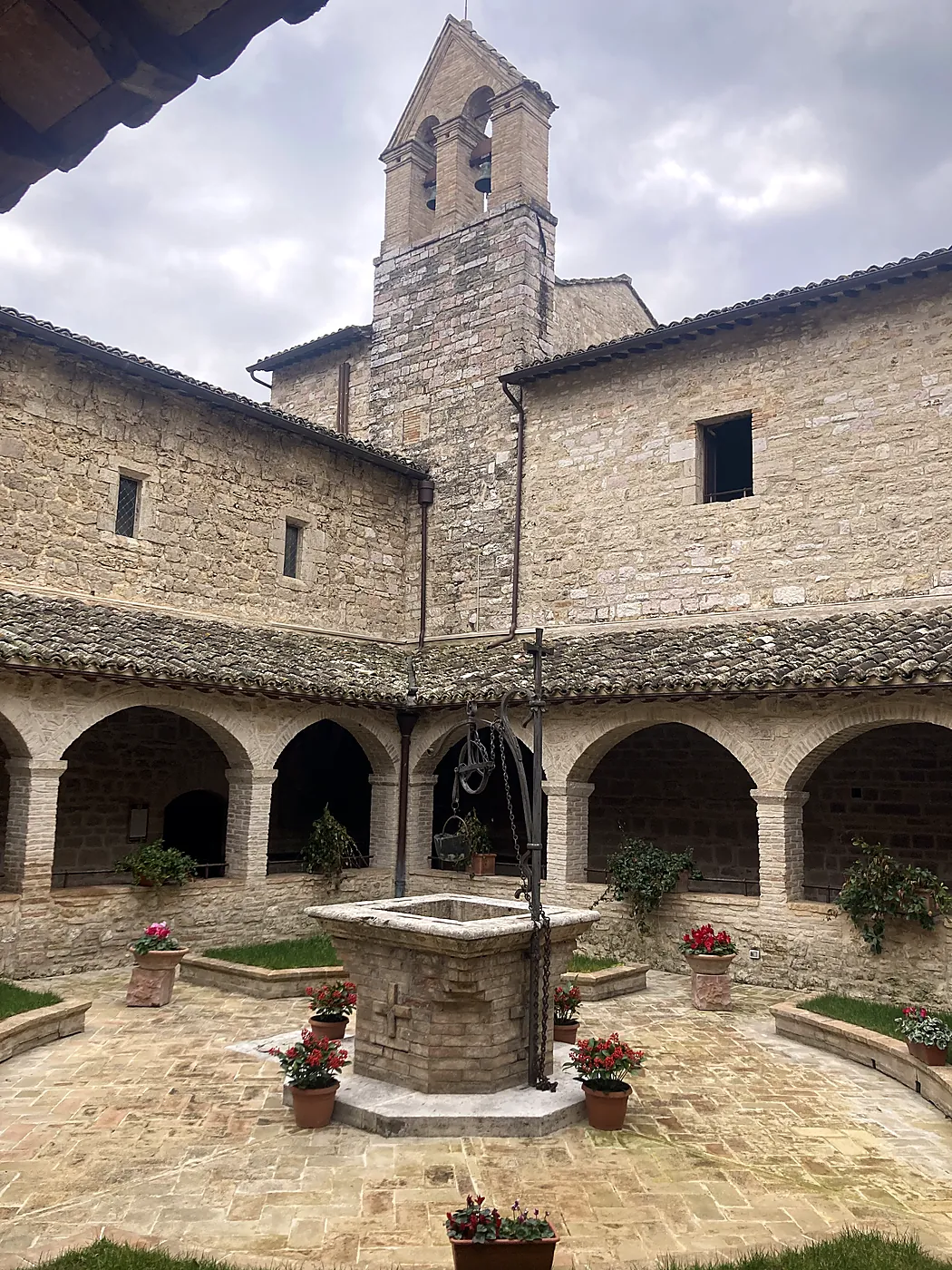 Il chiostro di un convento umbro, con il pozzo di pietra al centro, il campanile a vela e vasi di ciclamini rossi.