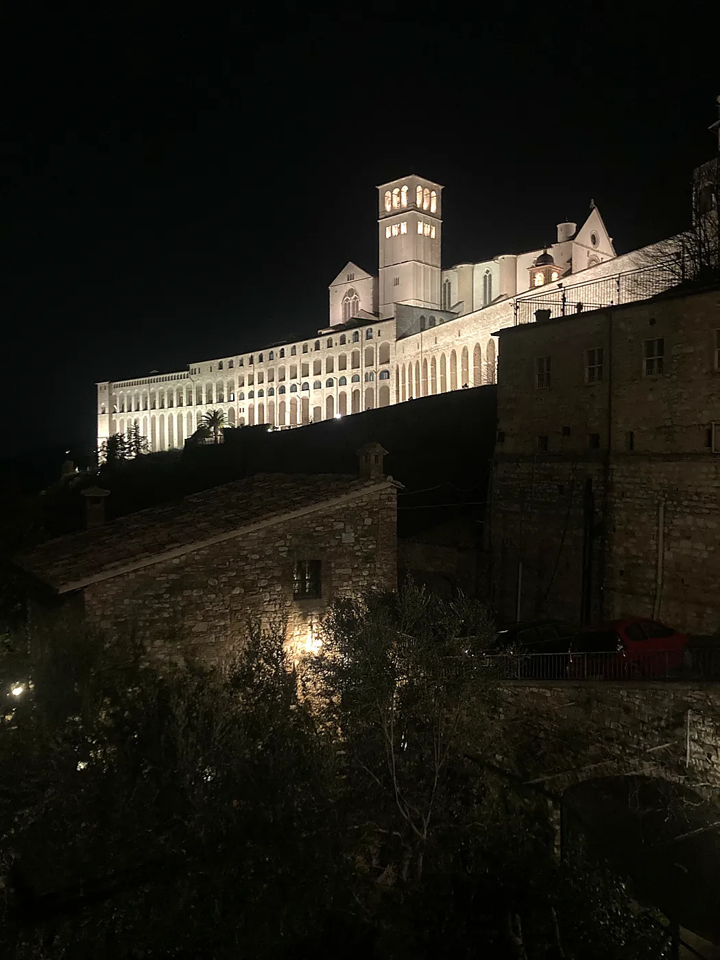 La basilica di San Francesco ad Assisi illuminata di notte, con il campanile e il lungo porticato del Sacro Convento sopra i tetti.