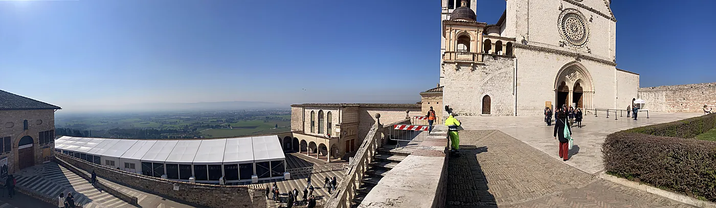 Veduta panoramica dal sagrato della basilica di San Francesco ad Assisi, con la facciata della chiesa superiore e la pianura umbra nella foschia.