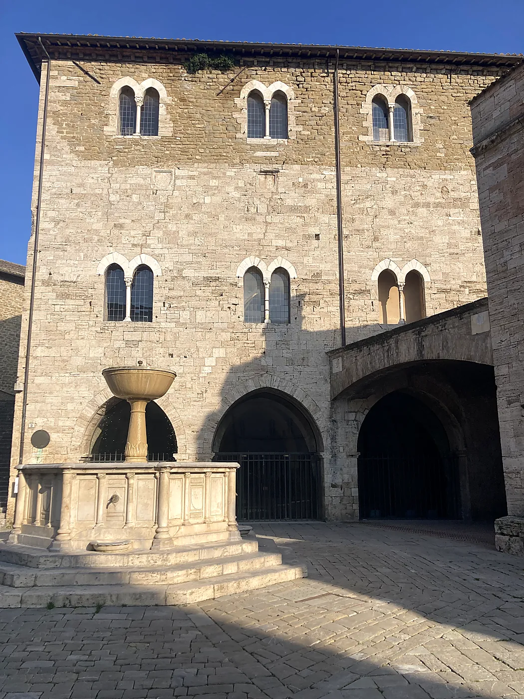 Il Palazzo dei Consoli e la fontana di piazza Silvestri a Bevagna, con le bifore gotiche nella luce del pomeriggio.