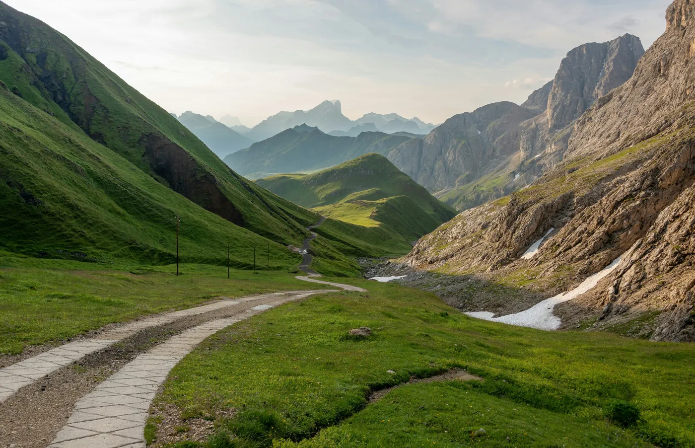 Un sentiero lastricato tra pascoli verdi in una valle di montagna, con pareti di roccia chiara e cime in lontananza