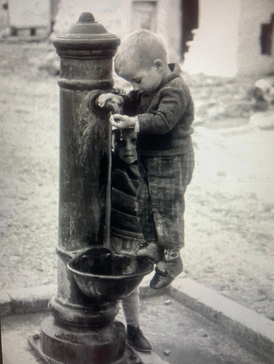Due bambini piccolissimi si aiutano a bere dal getto di una fontanella di ghisa